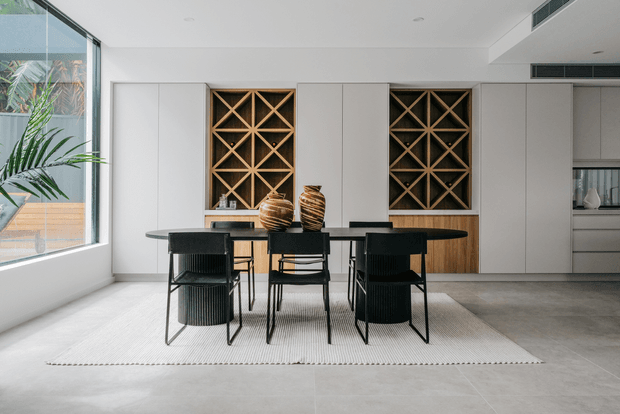 A bright contemporary dining area with a long dark dining table and black woven chairs, positioned on a large light rug. The room features pale grey built-in cabinetry, integrated timber wine racks with geometric lattice detailing, a warm timber lower cabinet section, concrete-look flooring, and large floor-to-ceiling glazing on the left. Decorative ceramic vessels sit centrally on the table.