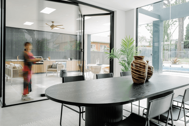 A bright contemporary dining area with a large dark oval dining table, black chairs, and a striped sculptural vase. Full-height black-framed glazing and internal glass partitions connect the dining space to an open kitchen and living area, with a pool and landscaped outdoor setting visible beyond. Light grey flooring, pale walls, vertical ribbed detailing, and indoor palms create a refined, airy interior.