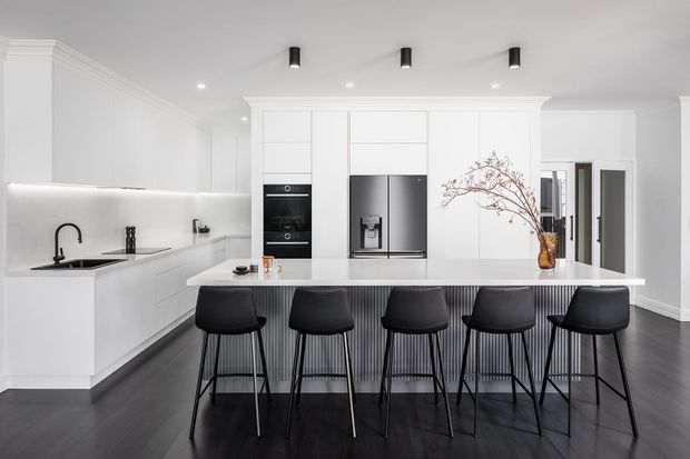 Bright contemporary open-plan kitchen featuring extensive white cabinetry, integrated black appliances, a long island with five black bar stools, a white benchtop, dark timber-look flooring, and subtle black lighting and tapware. Decorative branches add a small natural accent.