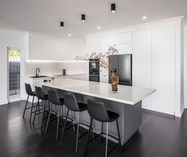 A bright contemporary kitchen with a large white island and integrated seating for five, white full-height cabinetry, a dark vertical fluted island base, dark timber flooring, and concealed appliances. Black pendant-style cylindrical downlights and recessed lighting provide contrast, while a vase of branches forms the island centrepiece.