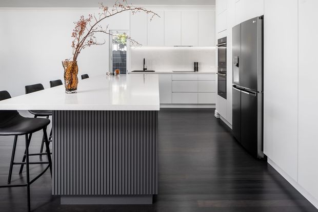 A contemporary kitchen with a large white island featuring a vertically fluted dark base and integrated seating. White handleless cabinetry, a white stone benchtop and splashback, dark timber-look flooring, black appliances and fixtures, and a simple branch arrangement create a clean, high-contrast interior.