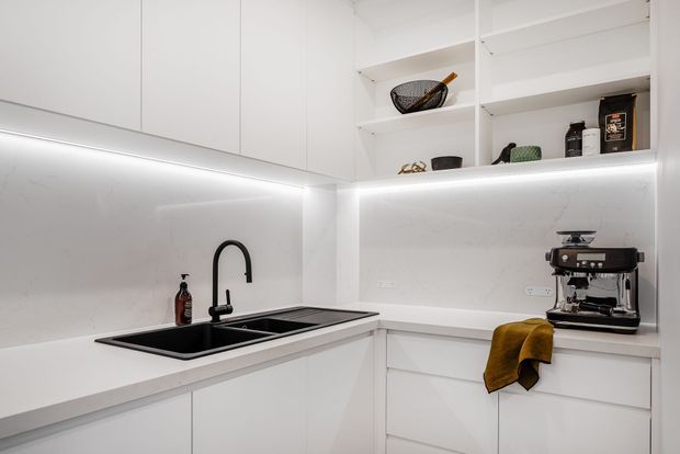 Bright, compact contemporary kitchen with white flat-panel cabinetry, a pale stone benchtop and splashback, integrated under-cabinet lighting, an undermount double sink with matte black gooseneck tap, open shelving, and a coffee machine on the corner counter. Mustard textile and dark fixtures provide restrained contrast.