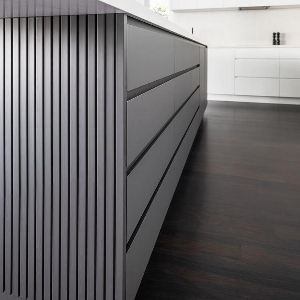 Close-up view of a contemporary kitchen island featuring dark grey, handleless drawer cabinetry and a vertically fluted side panel. The island has a light stone benchtop, set against white minimalist cabinetry and dark timber flooring.