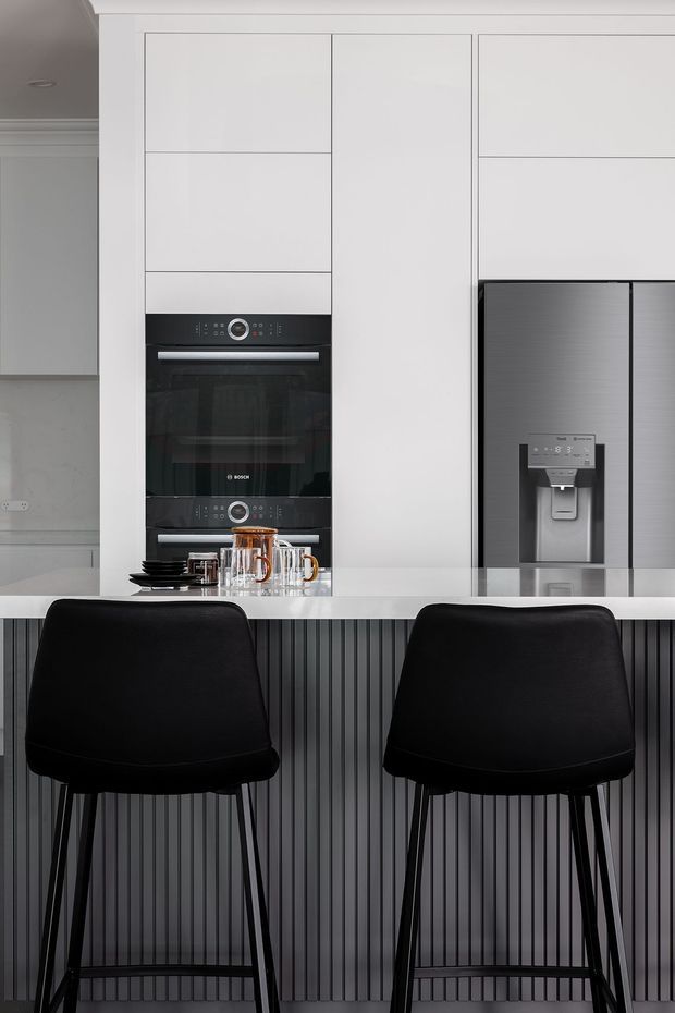 A contemporary kitchen with a white full-height cabinetry wall, integrated black Bosch ovens, and a large stainless-steel French-door refrigerator. A white stone-look island benchtop is paired with a dark vertically fluted island front and two black bar stools. Small coffee and tableware items sit on the benchtop.