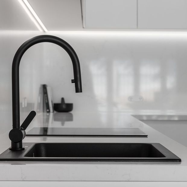 Close-up of a sleek contemporary kitchen sink and matte-black gooseneck tap set into a white benchtop. The black undermount sink, white glossy splashback, pale cabinetry, integrated cooktop and minimal linear lighting create a clean monochromatic composition.