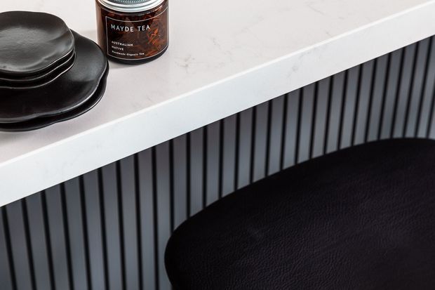 Close-up detail of a contemporary kitchen or cabinetry composition featuring a white marble-look benchtop, dark vertically fluted cabinetry, and a black leather-upholstered stool. Matte black plates and a tea jar sit on the benchtop.