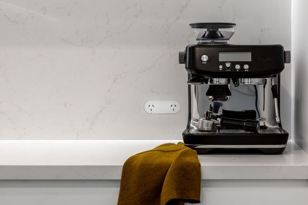 Close-up of a black espresso machine with an integrated grinder positioned on a white stone kitchen benchtop. The backsplash has subtle grey veining, with a mustard-yellow cloth draped over the front edge of the counter and a white power outlet visible behind the appliance.