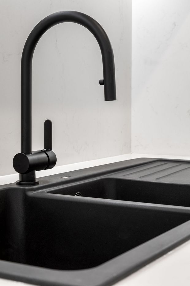 Close-up product-style image of a matte black gooseneck kitchen mixer tap mounted beside a deep black double-bowl sink with integrated draining grooves. The sink is set into a pale stone or marble-look benchtop and backsplash, creating a monochromatic, minimalist kitchen detail.