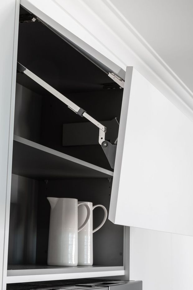 Close-up of a modern kitchen wall cabinet with a white lift-up door, dark interior shelving, and visible metal hinge hardware. Two white ceramic pitchers are stored inside, with part of a cooktop visible below.