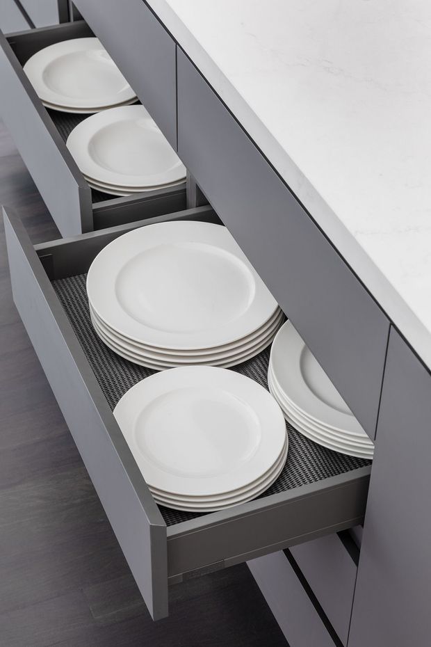 Close-up view of a modern kitchen drawer system with grey built-in cabinetry, a white stone-look benchtop, dark flooring, and neatly organised stacks of white plates on textured drawer liners.