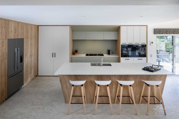 Bright contemporary kitchen with a large timber-clad island and four white bar stools, integrated dark refrigerator, white cabinetry, built-in double ovens, gas cooktop, pale benchtops, and a concrete-look floor. The room opens toward a glazed outdoor area and is finished with clean, streamlined joinery.