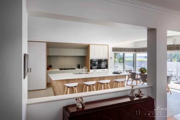 Bright contemporary open-plan kitchen viewed from an adjacent living or dining area. The kitchen features a large white island with timber-panelled base, four white bar stools, pale cabinetry, integrated appliances, a neutral splashback, and extensive glazing. A dining area and outdoor terrace are visible beyond, while a dark timber console occupies the foreground.