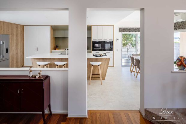 View through a hallway into a bright contemporary kitchen and dining area. The kitchen features light timber cabinetry, white cabinetry and benchtops, integrated appliances, a peninsula with stools, and pale stone-look flooring. Warm timber flooring and a dark timber console furnish the foreground, while a dining table and glazed doors are visible beyond.