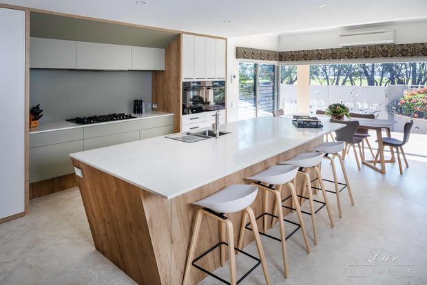 Bright open-plan kitchen and dining area with a large white island and integrated sink, timber-faced cabinetry, pale upper cupboards, and seating for several people on light timber stools. The kitchen opens to a glazed dining and outdoor area, with patterned clerestory-style window treatment and abundant natural light.