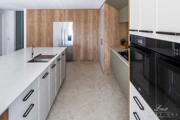 A contemporary galley-style kitchen with a large white island and integrated sink, white cabinetry, black appliances and hardware, warm wood-look wall cabinetry, and a light stone-effect floor. The space is bright, streamlined, and highly functional, with a stainless-steel refrigerator and built-in ovens visible.