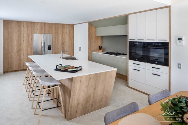 Bright contemporary open-plan kitchen featuring a large white island with integrated seating, light timber cabinetry and panels, white built-in cabinetry, and black integrated appliances. A dining area is visible in the foreground.