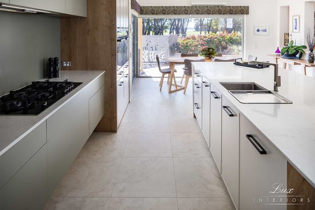 Bright contemporary kitchen viewed along a central walkway, with white flat-panel cabinetry, a large white island with integrated sink, black handles and fixtures, a dark stone or composite benchtop on the cooking run, and a full-height timber feature panel. Large rear glazing opens the kitchen toward a dining area and landscaped garden.