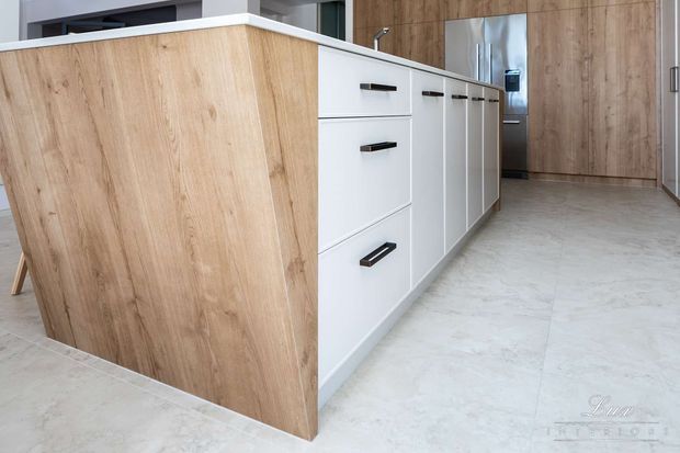Contemporary kitchen island with a light timber-clad end panel, white cabinetry and dark linear handles. The kitchen features a pale stone-look tiled floor, a white benchtop, and tall timber cabinetry in the background, creating a clean, restrained and warm modern interior.