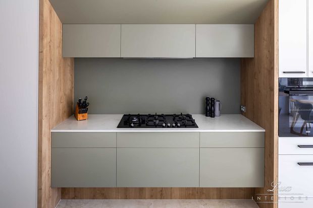 Compact contemporary kitchen with a streamlined grey and white built-in cabinet layout, white benchtop, dark grey splashback, integrated gas cooktop, and warm timber side panels framing the cooking zone. Additional white cabinetry and an oven are visible at the right edge.