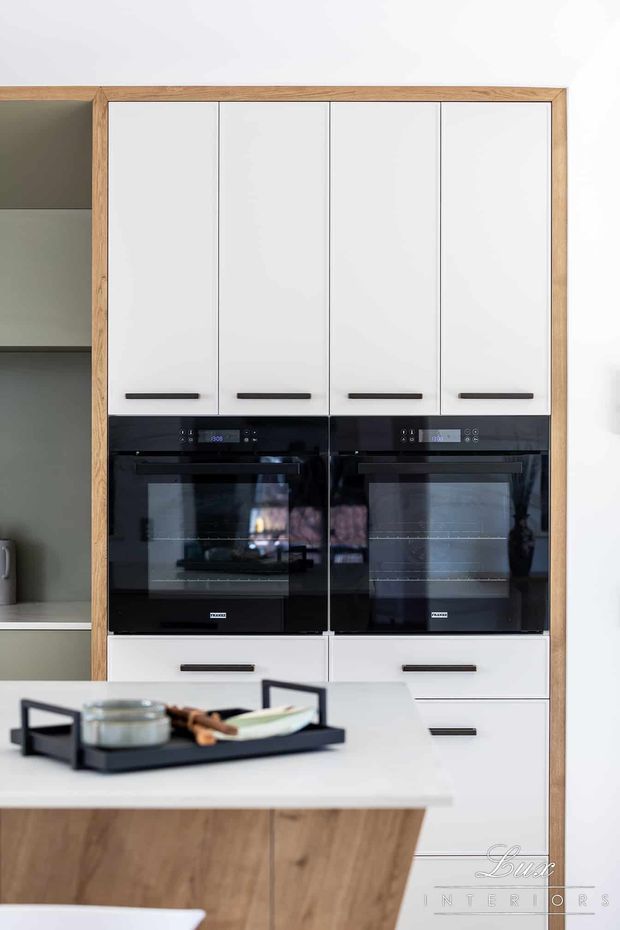 A contemporary kitchen featuring a tall bank of white handle-front cabinetry with integrated black double ovens, framed by warm natural timber. A white island with a timber-clad side occupies the foreground, with a dark serving tray on top.