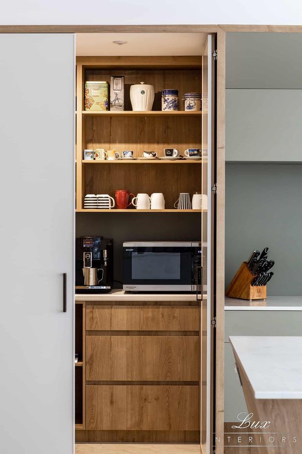 A tall built-in kitchen appliance and storage pantry with open shelves displaying crockery and decorative vessels, integrated microwave and coffee machine, and lower timber drawers behind a partially open white door. Adjacent pale cabinetry and a stone island edge are visible.