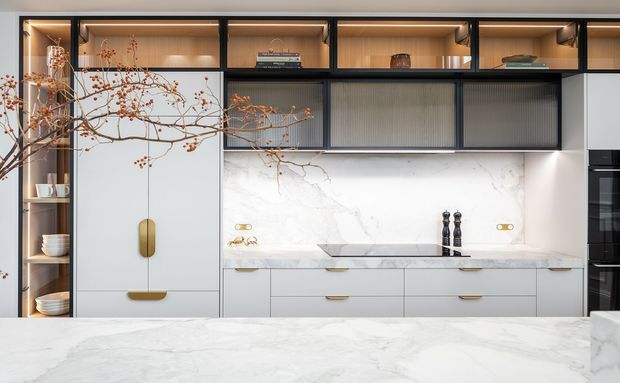Contemporary kitchen with a large pale marble island in the foreground, white marble splashback and benchtops, integrated white cabinetry with brass-toned handles, and dark-framed glass-fronted overhead cabinetry. Warm internal cabinet lighting and open shelving display ceramics and decorative objects, while a sculptural branch adds an organic accent.
