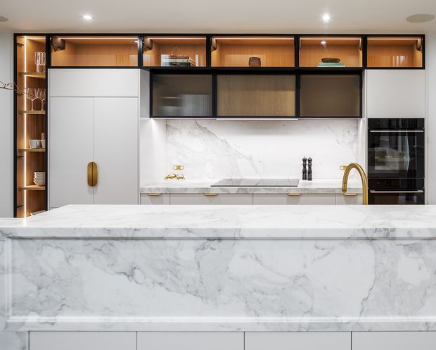 A contemporary kitchen featuring a large veined marble island with waterfall-style detailing, white full-height cabinetry, warm illuminated timber display niches, dark-framed upper joinery, integrated appliances, and a brass gooseneck tap. The palette combines white stone, pale cabinetry, black accents, warm timber, and brass hardware.