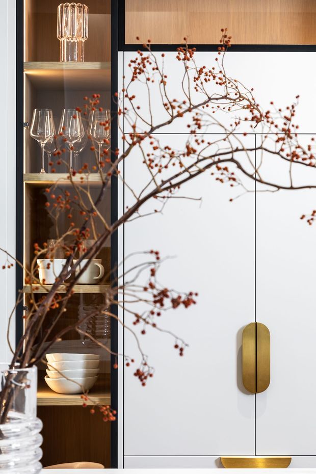 Close-up interior detail of a refined built-in storage and display cabinet. Warm wood open shelving holds glassware and ceramics, while adjacent white cabinetry has slim black framing and sculptural brass-gold handles. A decorative branch in a glass vase crosses the foreground, and integrated shelf lighting adds warmth.