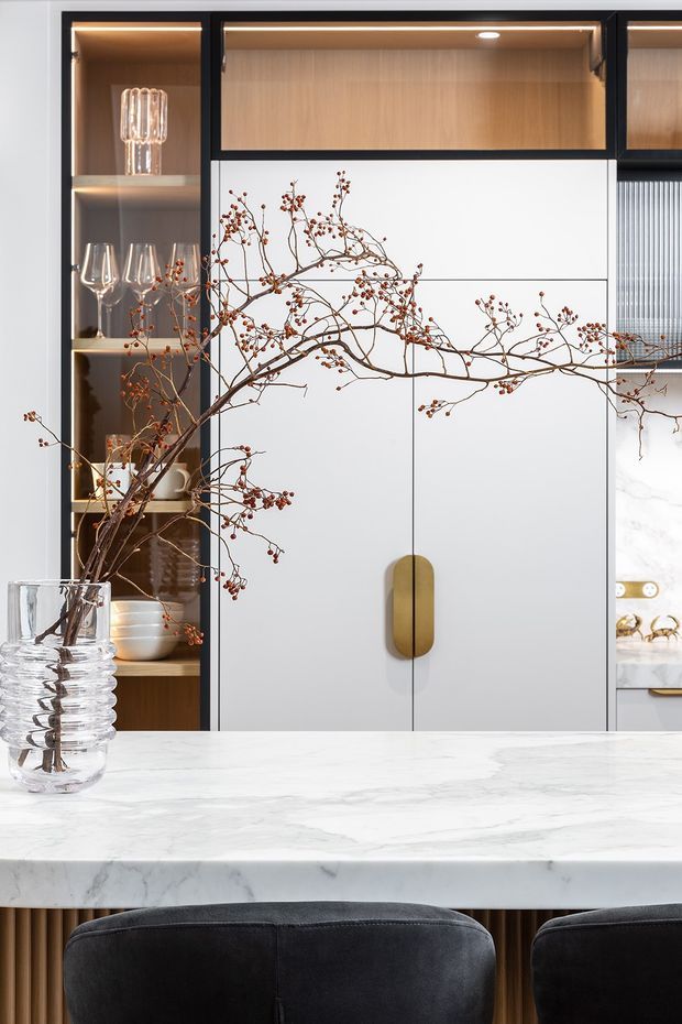 A refined contemporary kitchen featuring a large white marble island with rounded edge, dark upholstered stools, integrated white cabinetry with a brushed brass handle, warm timber display shelving, black-framed joinery, and softly illuminated glassware niches. A sculptural branch arrangement in a clear glass vase adds an organic decorative element.