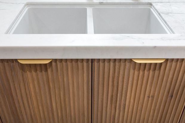 Close-up detail of a double-basin kitchen sink set into a white marble-look benchtop, with warm vertically fluted timber cabinetry and brass-toned recessed pulls below.