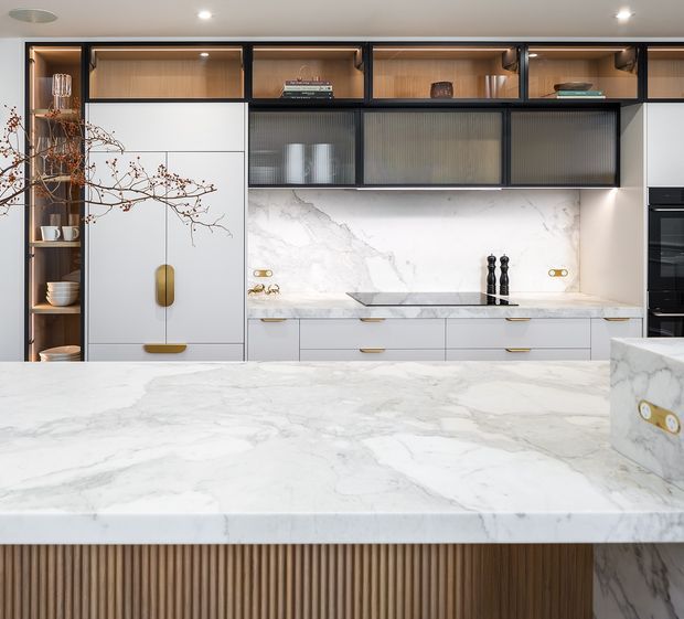 A contemporary kitchen with a large white marble island featuring a vertically fluted timber base, white marble splashback and benchtops, integrated white cabinetry with brass handles, warm timber display shelving, and dark-framed ribbed-glass overhead cabinets. Built-in appliances are visible at the right.