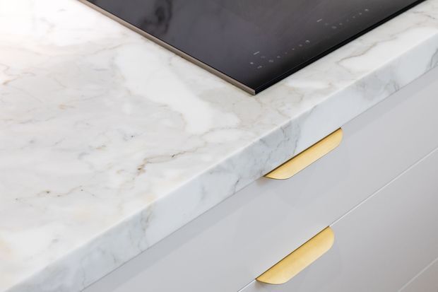 Close-up detail of a contemporary kitchen or cabinetry installation, showing a white veined marble benchtop, black induction cooktop, white drawer fronts, and brushed gold curved handles.