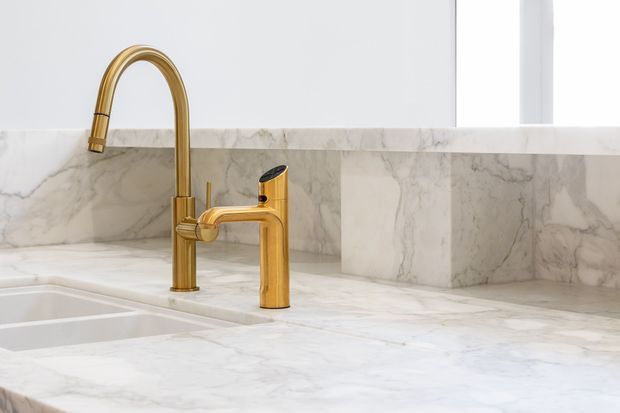 Close-up view of a refined kitchen sink area featuring a white marble countertop and matching marble splashback, with a sculptural brushed brass gooseneck kitchen tap and separate brass mixer fixture. Bright natural light enters from a nearby window.