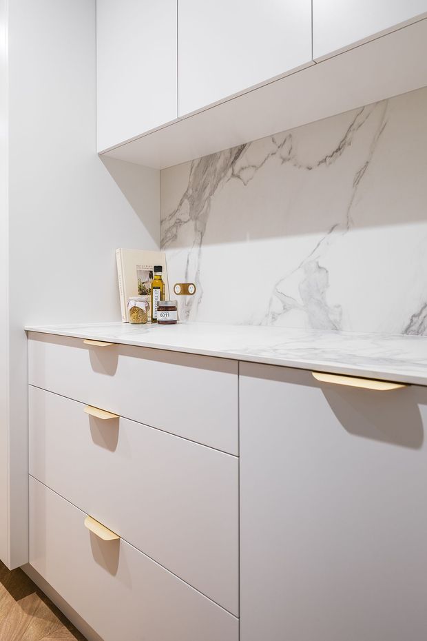 Close-up of a refined, minimalist kitchen run featuring pale matte cabinetry with integrated drawer fronts and sculptural brass-gold handles. A white marble-look stone benchtop and matching full-height veined splashback sit beneath sleek overhead cupboards. The composition is bright, clean, and highly streamlined.