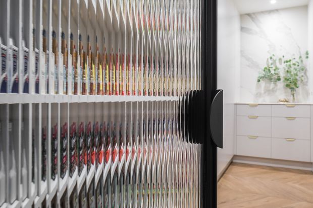 A contemporary pantry or storage room viewed through a partially open tall cabinet door. The foreground is dominated by extensive vertical shelving filled with pantry goods, while the background shows white built-in cabinetry with slim brass pulls, a marble-look wall panel, greenery, recessed lighting, and a light herringbone timber floor.