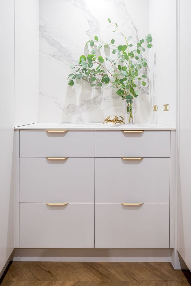 A compact, refined built-in storage cabinet with six pale drawers and curved brass-gold pulls. The cabinet has a light stone or marble-look benchtop and a dramatic white marble-look backsplash with grey veining. A clear glass vase with leafy branches and a small decorative object sit on the counter. Herringbone timber flooring and white walls complete the bright, elegant interior.