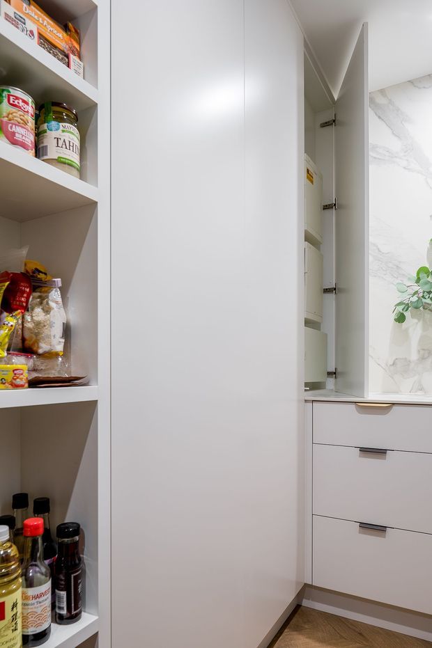 A compact, highly organised pantry with full-height white cabinetry, open shelving stocked with pantry goods, and integrated drawers. The cabinetry has a clean minimalist finish, with a marble-look wall surface visible at right and warm timber-look flooring below.
