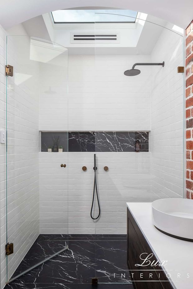 Contemporary walk-in bathroom shower featuring white horizontal wall tiles, dramatic black marble-look floor and feature band tiles, matte dark shower fittings, frameless glass enclosure, a white vessel basin on dark timber cabinetry, exposed red brick, and a skylight providing natural light.