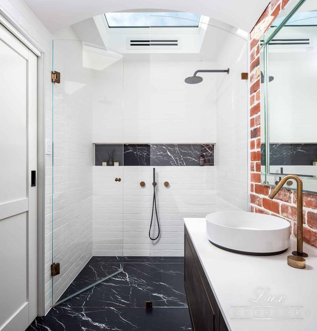 Bright contemporary bathroom featuring a frameless glass walk-in shower, white tiled walls, a skylight, black marble-look floor and horizontal feature band, exposed red brick, and a floating dark vanity with a round vessel basin and brushed brass tapware.