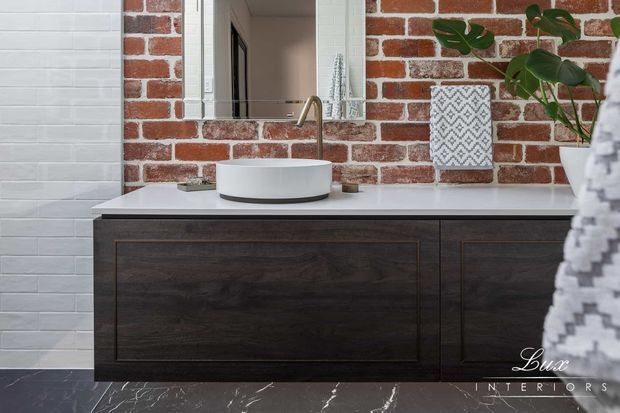 A contemporary bathroom vanity with a dark timber floating cabinet, white vessel basin, and brass-toned tapware. The walls combine exposed red brick with white rectangular wall tiles, while a framed mirror, patterned wall art, and a potted plant add decorative detail. Dark marble-look flooring completes the scheme.
