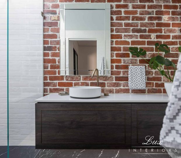 A compact contemporary bathroom vanity featuring a dark timber floating cabinet, white vessel basin, brushed brass tapware, and a rectangular mirror. The main wall is exposed red brick, contrasted with white tiled wall panels and dark marble-look flooring. A patterned vase and indoor plant add decorative detail.
