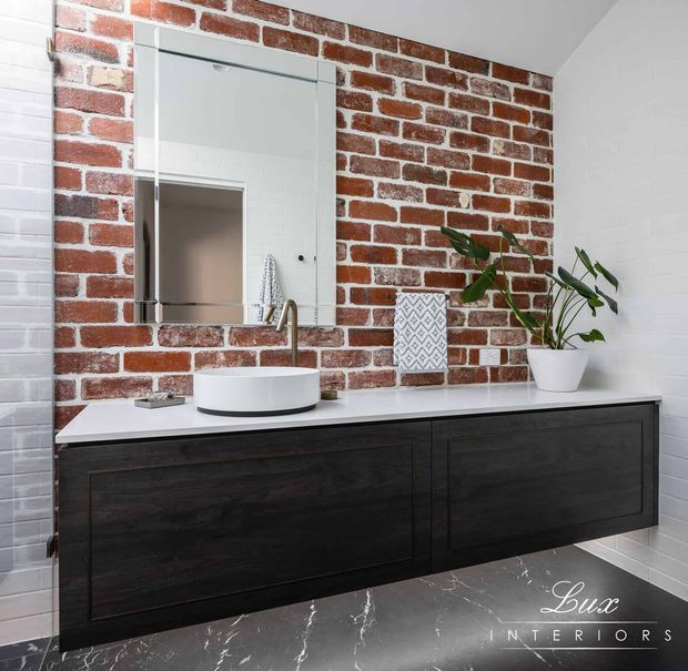 Contemporary bathroom vanity with a long floating dark timber-toned cabinet, white vessel basin, brushed brass tapware and white benchtop. A large frameless mirror is set against a red exposed-brick feature wall, complemented by white brick walls and dark marble-look floor tiles. Simple patterned and botanical accessories add contrast.