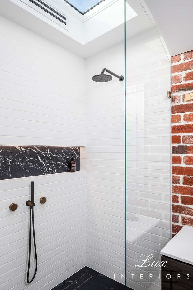 A bright contemporary walk-in shower with white rectangular wall tiles, a black marble-look feature niche and shower floor, dark metal shower fixtures, a frameless glass screen, skylight, and an exposed red brick wall detail.
