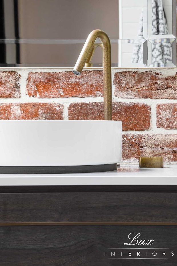 Close-up bathroom vanity featuring a white rectangular vessel basin on a dark timber vanity, a brushed brass gooseneck tap, exposed red brick backsplash, white countertop, and a mirrored wall with decorative bevelled mirror panels. The image has a refined industrial character.