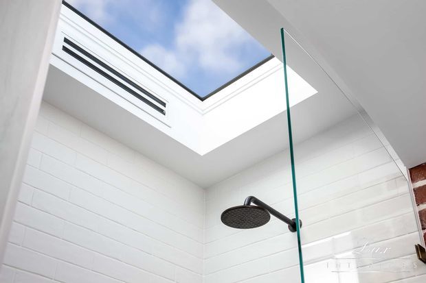Bright contemporary shower enclosure with white horizontal wall tiles, a large overhead rain shower, frameless glass, and a skylight above. A slim linear ventilation grille is visible near the ceiling, with a partial exposed brick edge at the right.