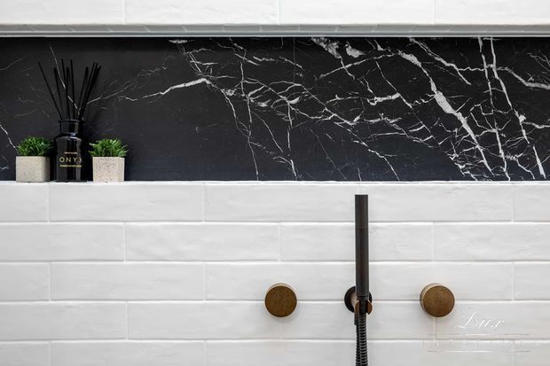 Close-up of a contemporary bathroom shower wall featuring white horizontally laid rectangular tiles, a dramatic black marble-look feature strip with white veining, and brushed brass-gold shower controls and fixtures. Small potted plants and a reed diffuser sit on the upper ledge.