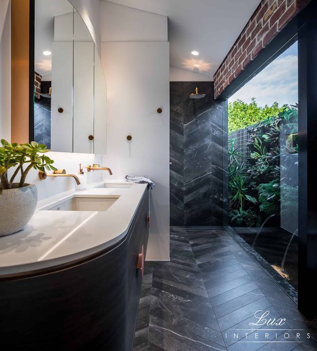 A sophisticated contemporary bathroom featuring a long white vanity with integrated basins, brushed brass tapware, mirrored cabinetry, dark timber joinery, and charcoal stone wall and floor tiles. A glass-enclosed shower opens toward a lush planted outdoor area, while a brick feature wall and black-framed glazing add texture and contrast.