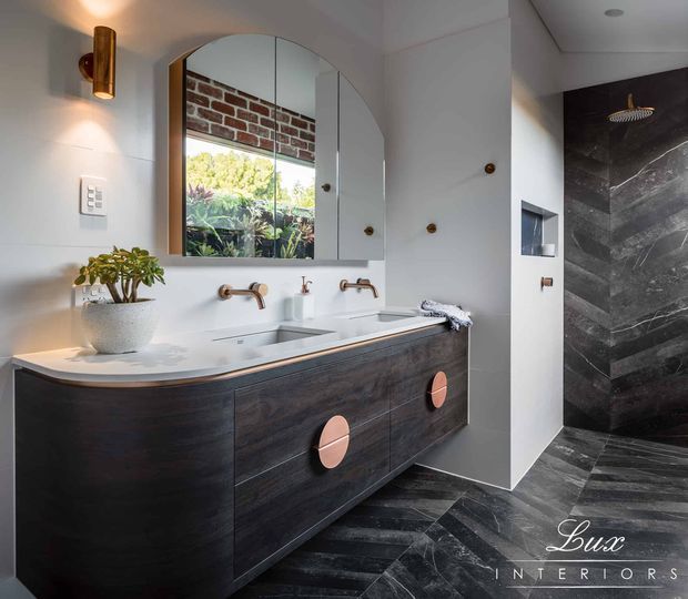 A refined contemporary bathroom featuring a large floating dark timber double vanity with a pale stone benchtop, twin basins and wall-mounted brushed metal taps. An arched mirrored cabinet spans the vanity, reflecting a brick wall and greenery. The room includes white tiled walls, a sculptural wall sconce, and a separate shower lined in dramatic dark veined stone with matching dark flooring. Warm copper-toned hardware provides contrast.