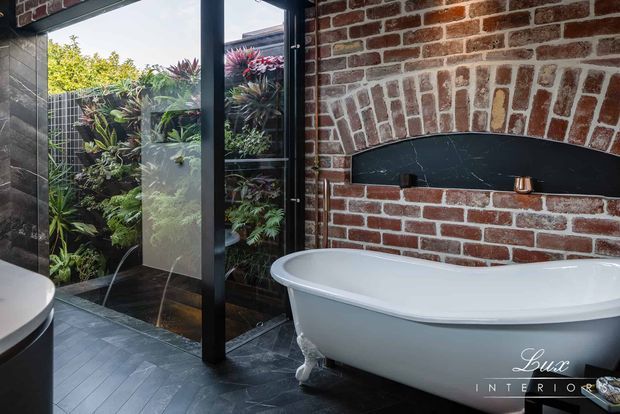 A luxurious bathroom featuring a freestanding white bathtub positioned against an exposed red brick wall with a distinctive arched black inset. Dark slate-like floor tiles, black-framed glazing, and a lush planted courtyard visible through the glass create a dramatic indoor-outdoor atmosphere.
