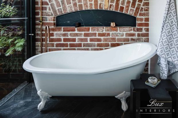 A refined bathroom featuring a white freestanding clawfoot bathtub set against an exposed red-brick wall with a curved black marble-look inset. Dark stone floor tiles, black-framed glazing, brass plumbing fixtures, and patterned textiles create a dramatic, vintage-inspired interior.